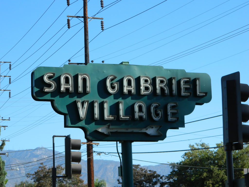 Prominent green sign with neon text that reads "San Gabriel Village". Under the text is an arrow pointing the way.